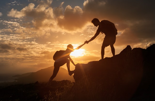 Male and female hikers climbing up mountain cliff and one of them giving helping hand. People helping and, team work concept.